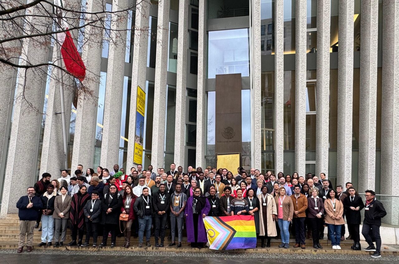 Imagen de un numeroso grupo de personas posando frente a un edificio, sosteniendo una pequeña bandera del Orgullo.