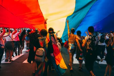 Crowd marching under a large Pride flag.